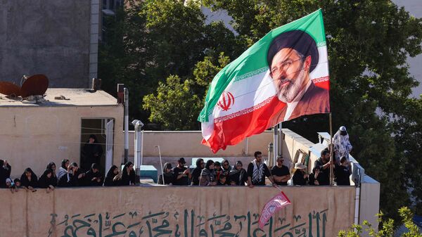 A flag of Iran bearing the image of Iran's current supreme leader Mojtaba Khamenei is seen on top of a building as people watch mourners take part in a funeral procession for Iran's slain supreme leader Ali Khamenei and members of his family in Mashhad on July 9, 2026. (Photo by ATTA KENARE / AFP) /